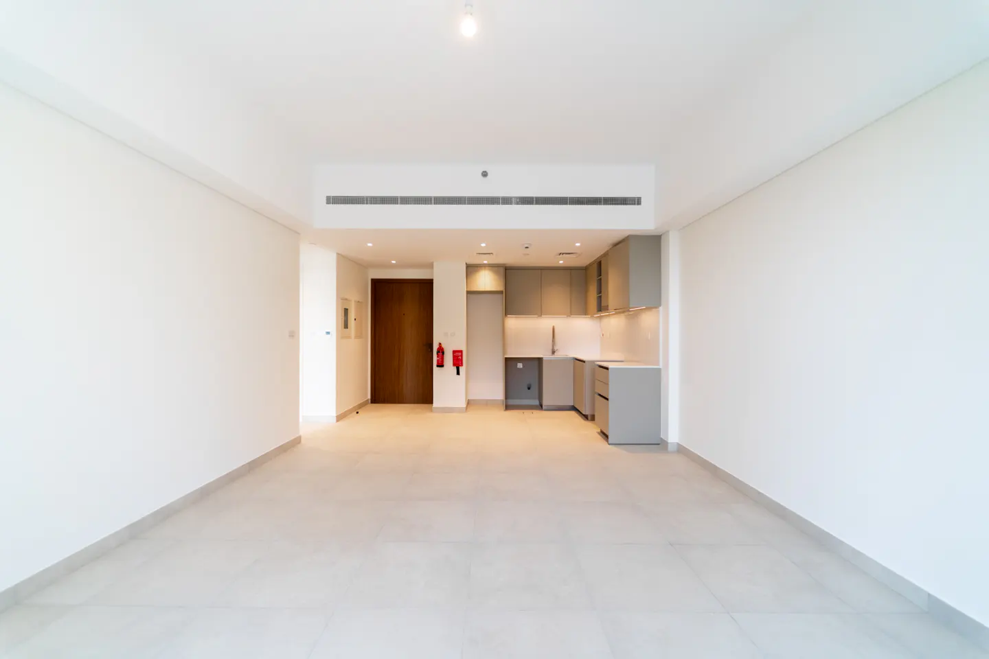 A bright, empty apartment with white walls and tile floors. A small kitchen area with gray cabinets is visible in the background.