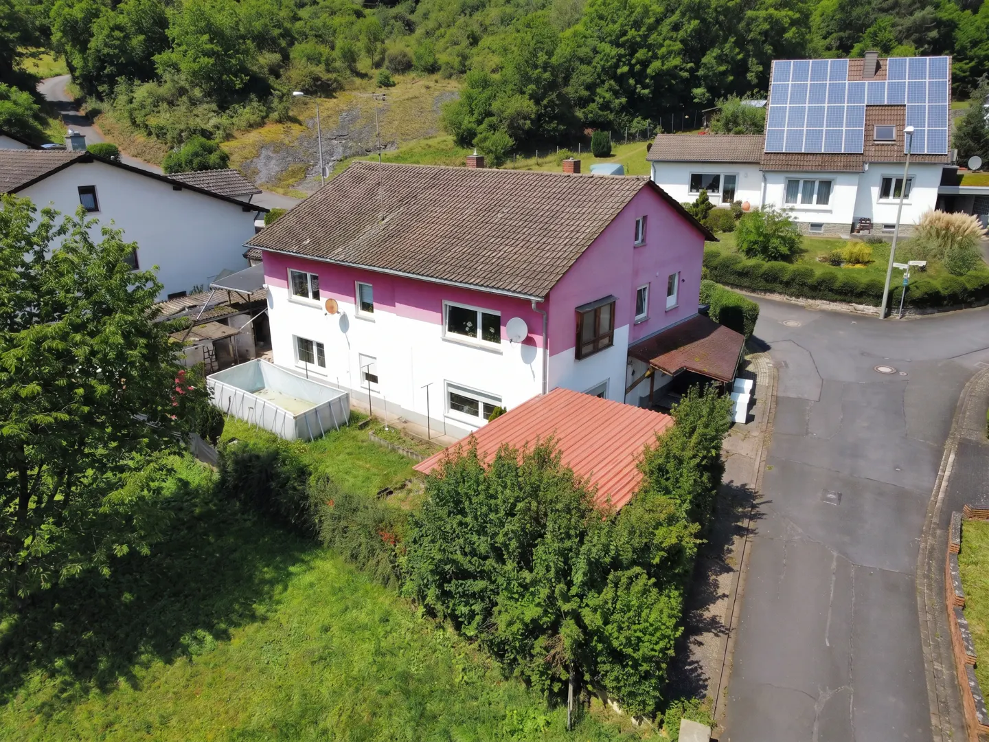 Aerial view of a two-story house with pink and white exterior, brown roof, and a pool in the backyard. Green trees surround the property.