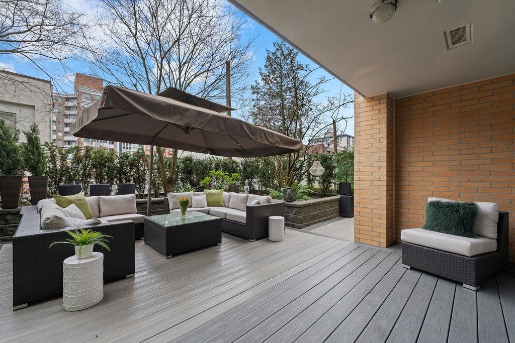 Outdoor patio with gray wood flooring, wicker furniture, and a brown umbrella. A brick wall and green plants add texture.