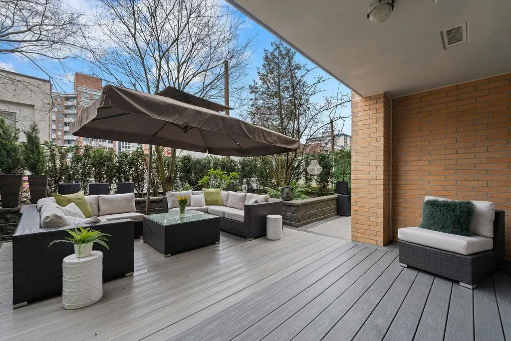 Outdoor patio with gray wood flooring, wicker furniture, and a brown umbrella. A brick wall and green plants add texture.