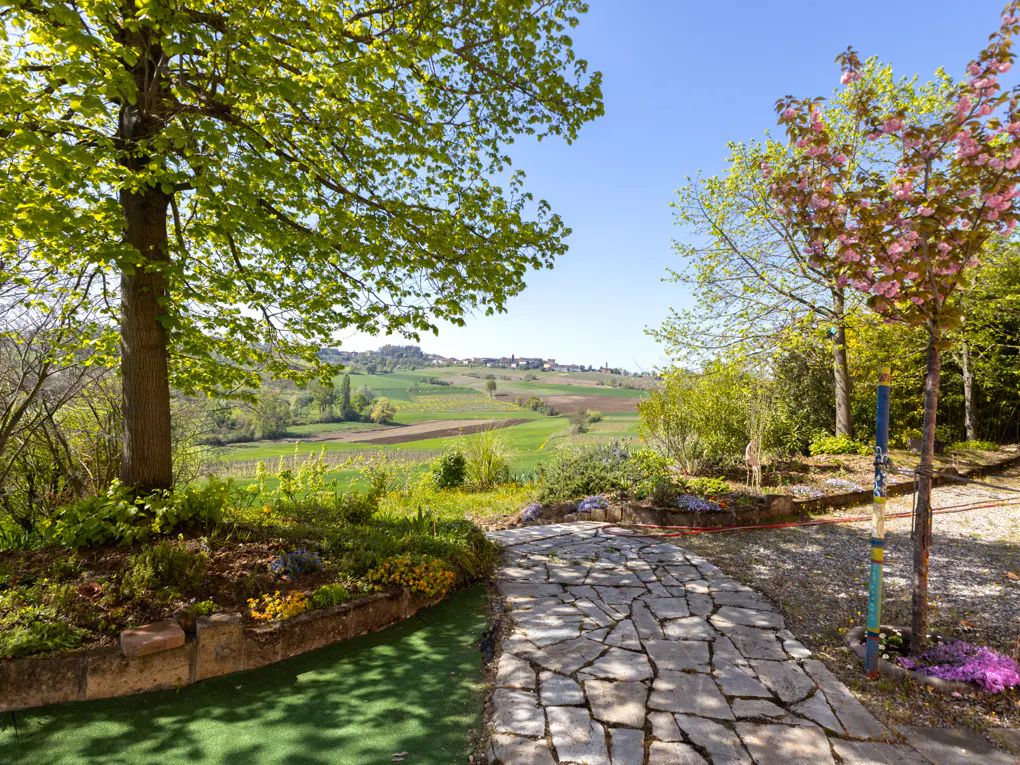 Stone path leads to a green, hilly landscape under a blue sky. Trees with green and pink blossoms frame the view.