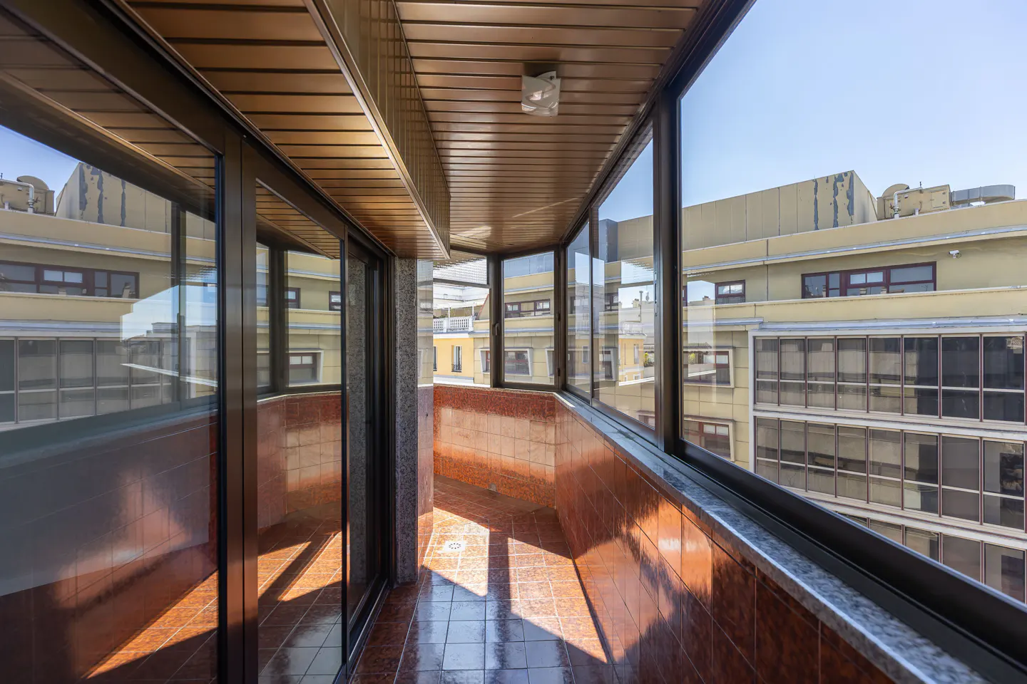 Enclosed balcony with brown tile floor and walls, dark-framed windows, and a wood-paneled ceiling, overlooking a building.
