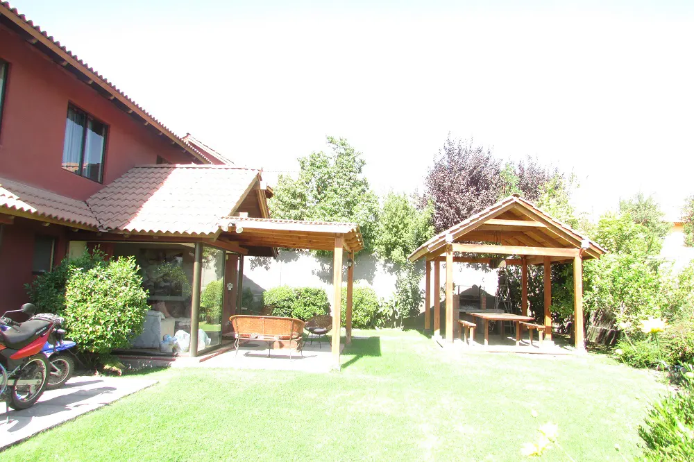 A backyard with a red house, green lawn, two wooden gazebos, and a motorcycle parked near the house.