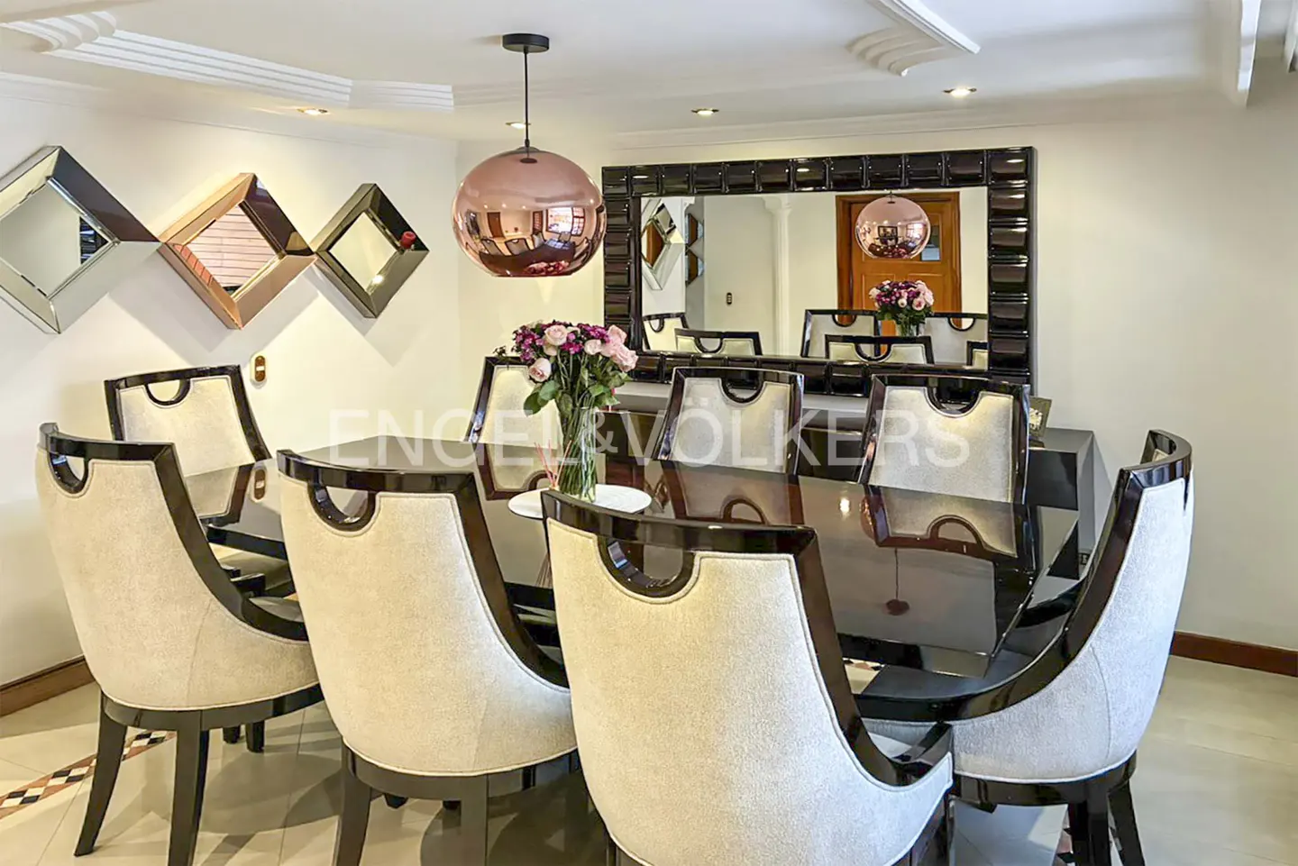 Modern dining room with a dark glossy table, cream upholstered chairs, a rose gold pendant light, and wall mirrors.
