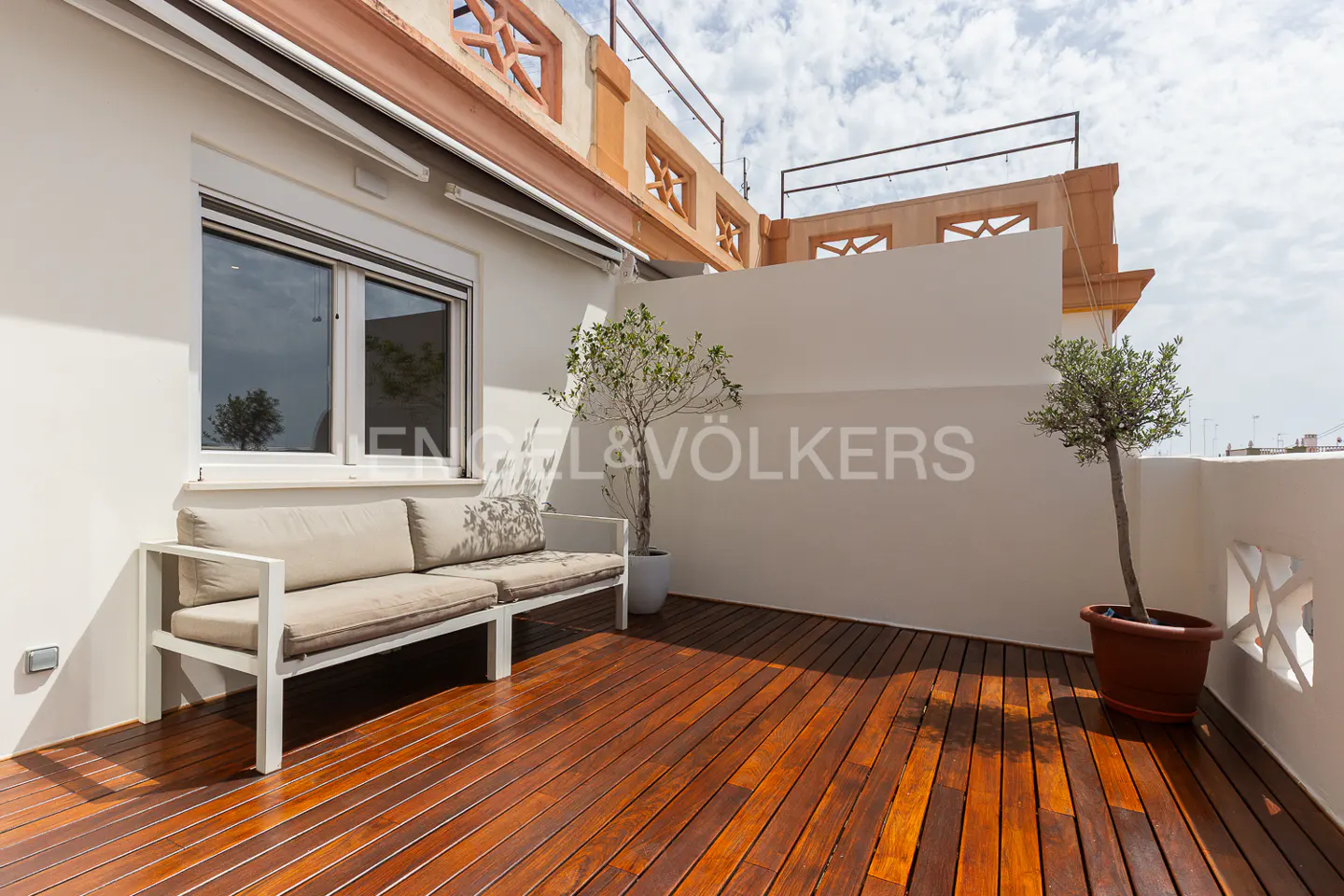 Outdoor patio with a wooden floor, a white couch, and two potted trees against a white wall.