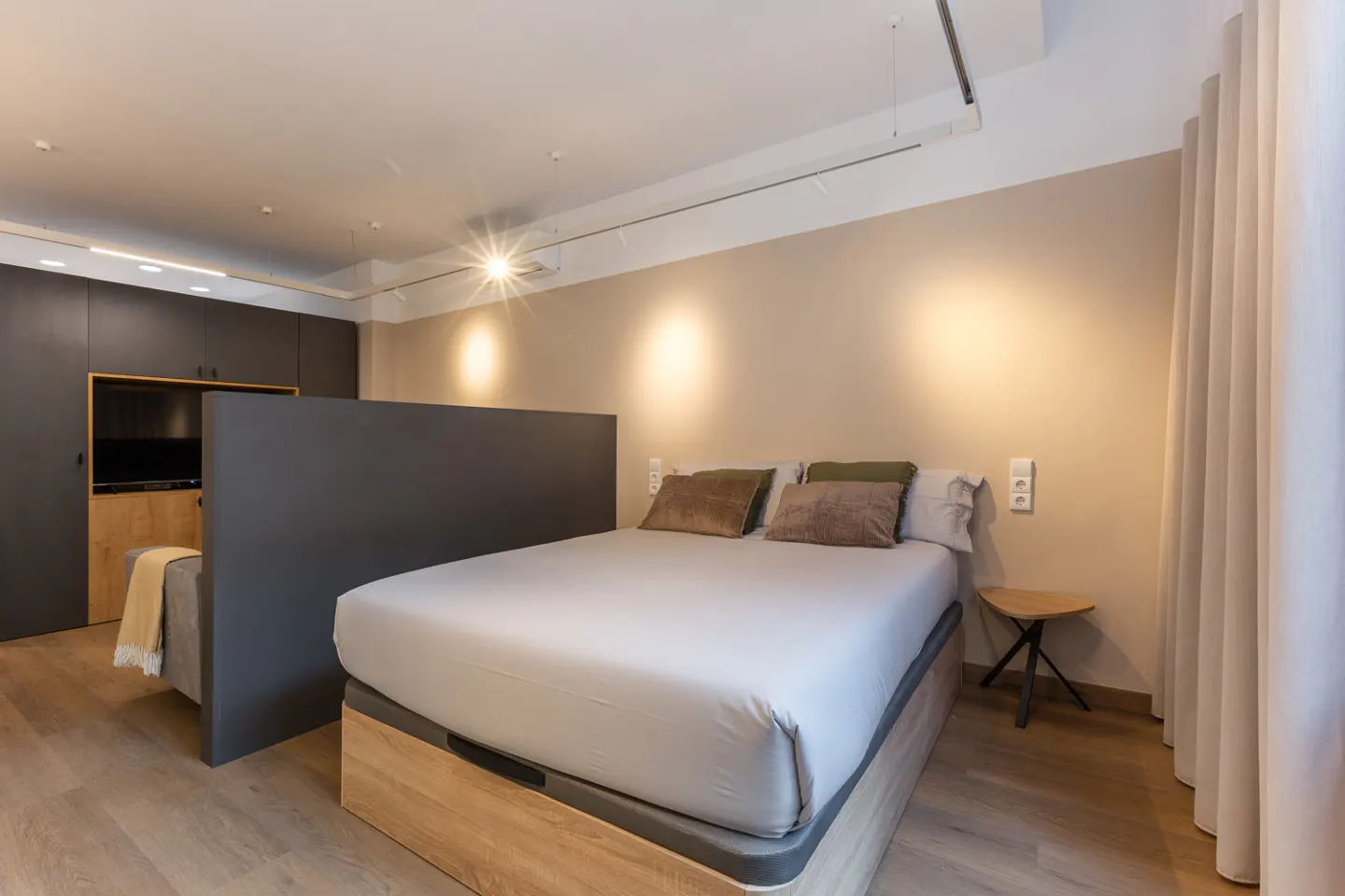 A modern bedroom with a gray bed frame, white sheets, and brown pillows. A gray partition separates the bed from a seating area with a TV.