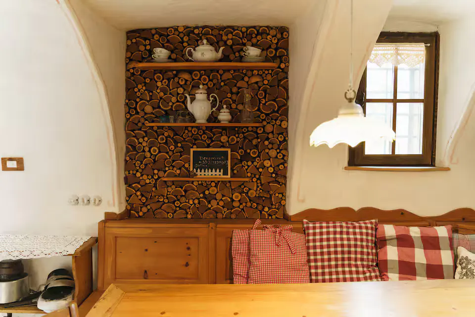 Cozy dining area with wood-paneled wall, shelves holding teacups, and a wooden bench with red and white checkered pillows. A window provides natural light.