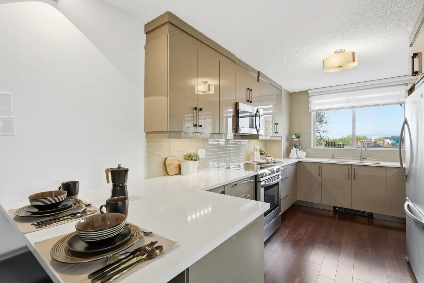 A modern kitchen with tan cabinets, stainless steel appliances, and white countertops. A table is set with plates and silverware.
