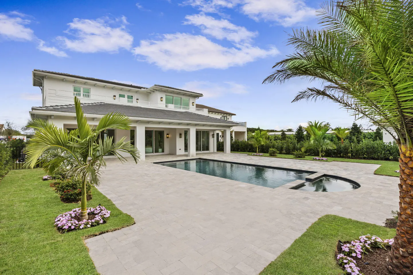 Backyard view of a two-story white house with a pool, patio, and palm trees under a blue sky with clouds.