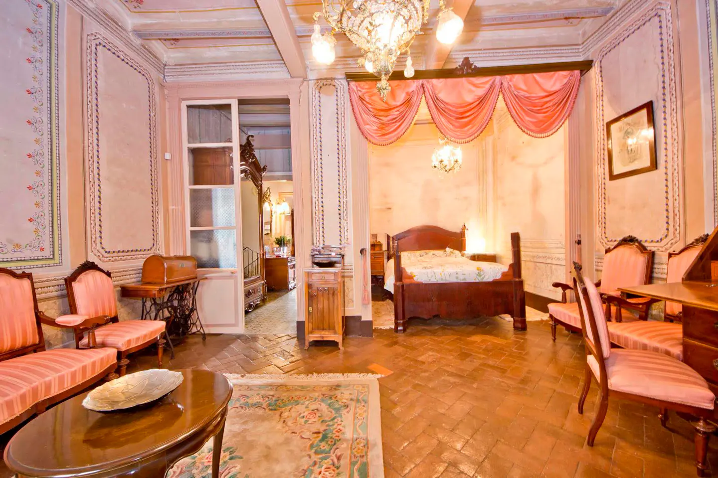 Ornate bedroom with pink walls, brick floors, and antique furniture. A bed with a pink canopy is visible through an archway.