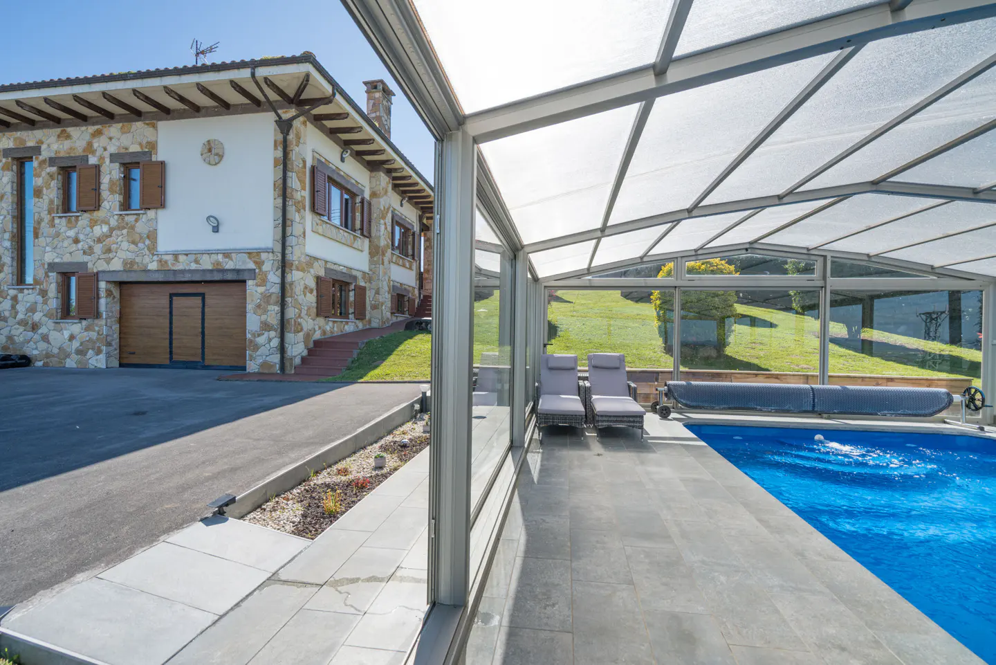 Exterior view of a stone house with a covered pool. Two lounge chairs sit beside the pool, with a green lawn in the background.