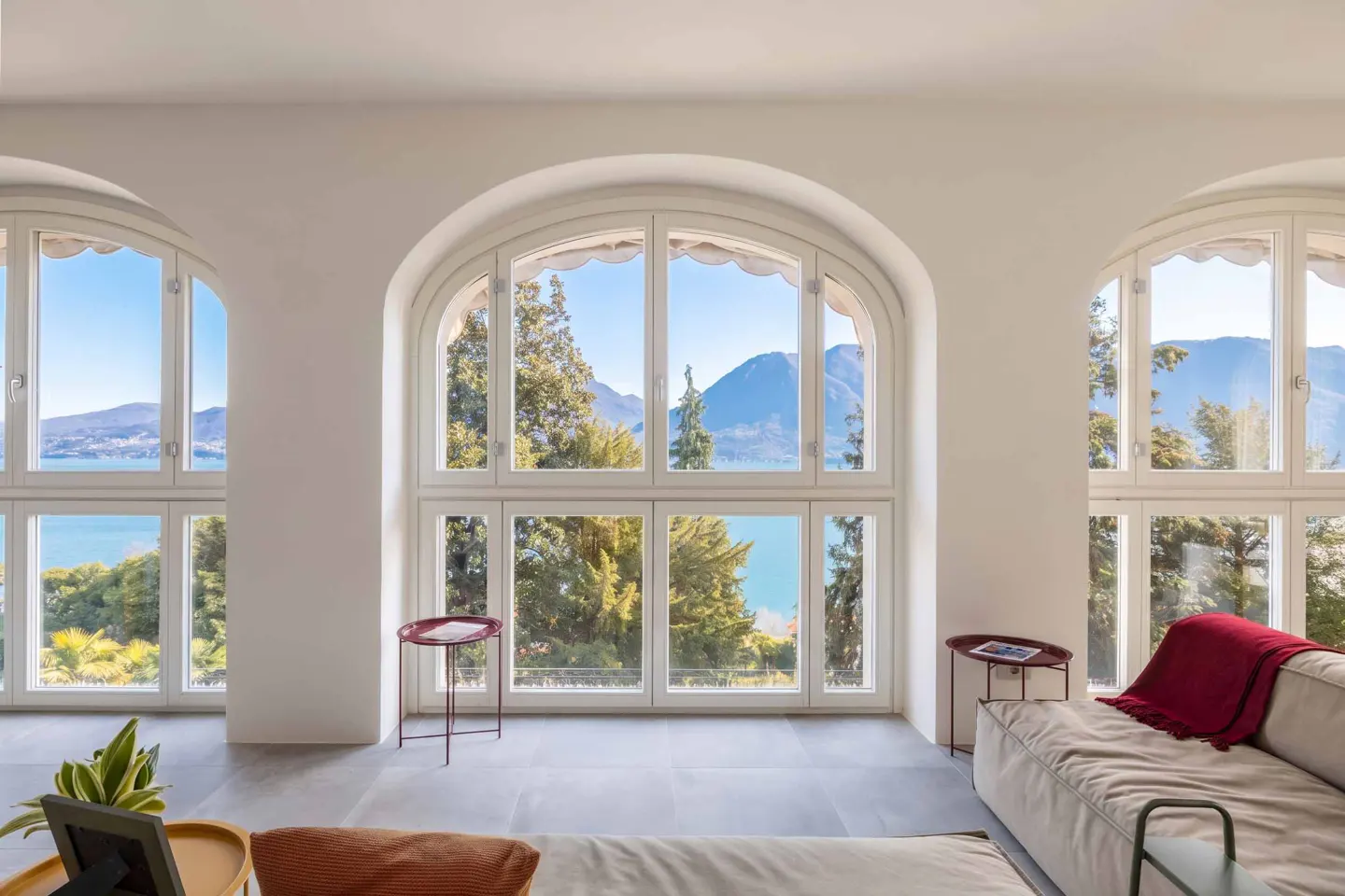 Bright room with three arched windows framing a lake view. A beige sofa with a red throw blanket sits near a small red table.