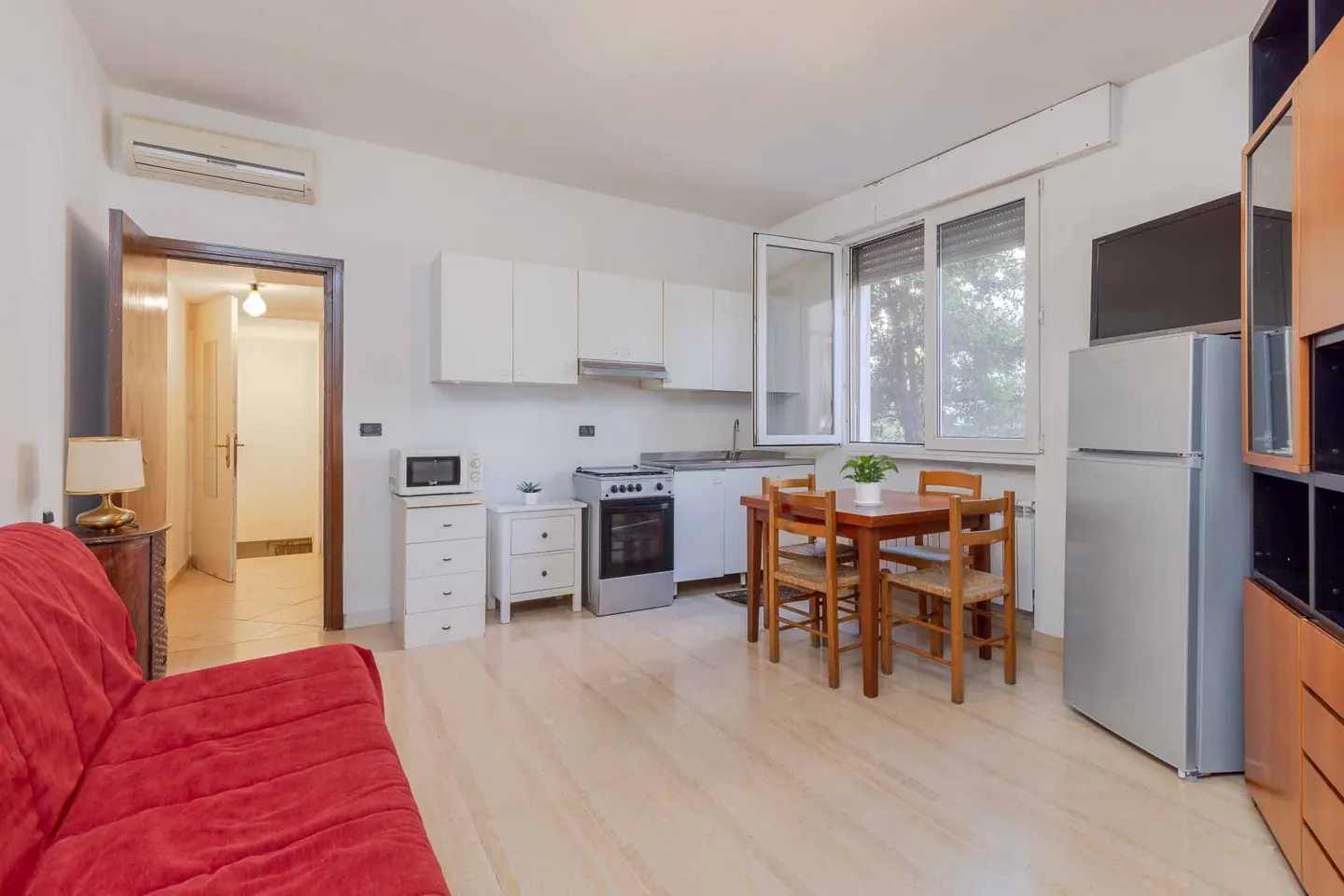 A bright studio apartment with a red sofa, a kitchen with white cabinets, and a wooden dining table.