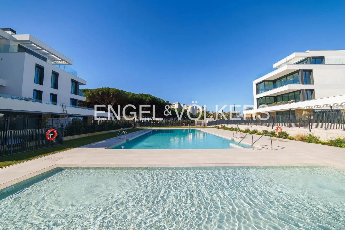 Outdoor pool area with modern white buildings on either side under a clear blue sky. The Engel & Völkers logo is visible.