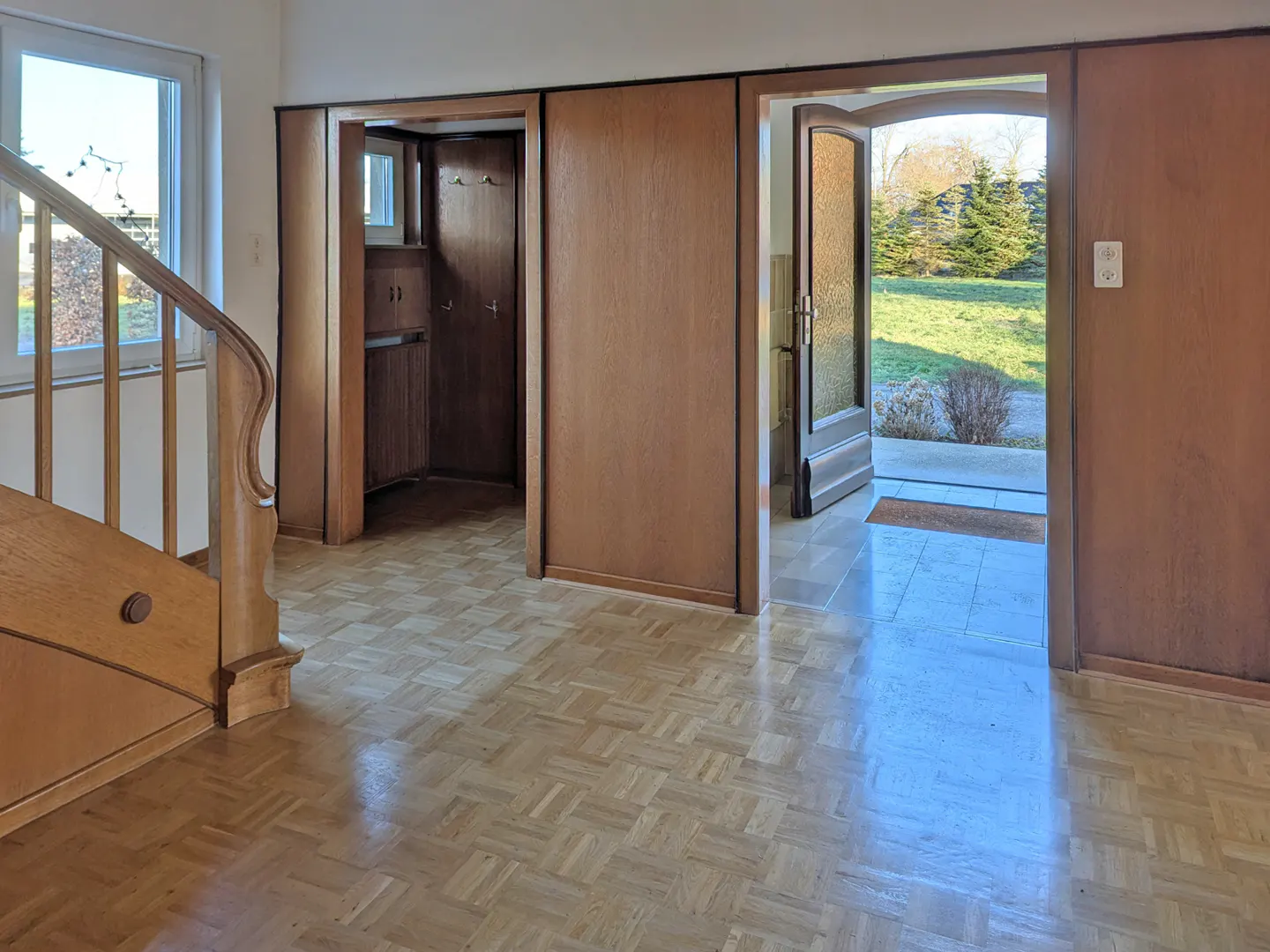 Interior view of a home's entryway with parquet floors, wood staircase, coat closet, and open door to the outside.