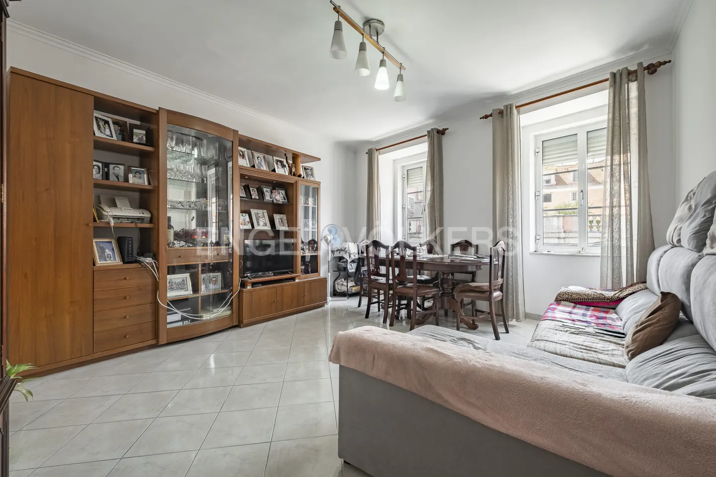 A living room with a large wooden cabinet, dining table, and gray sofa. Windows with curtains let in natural light.