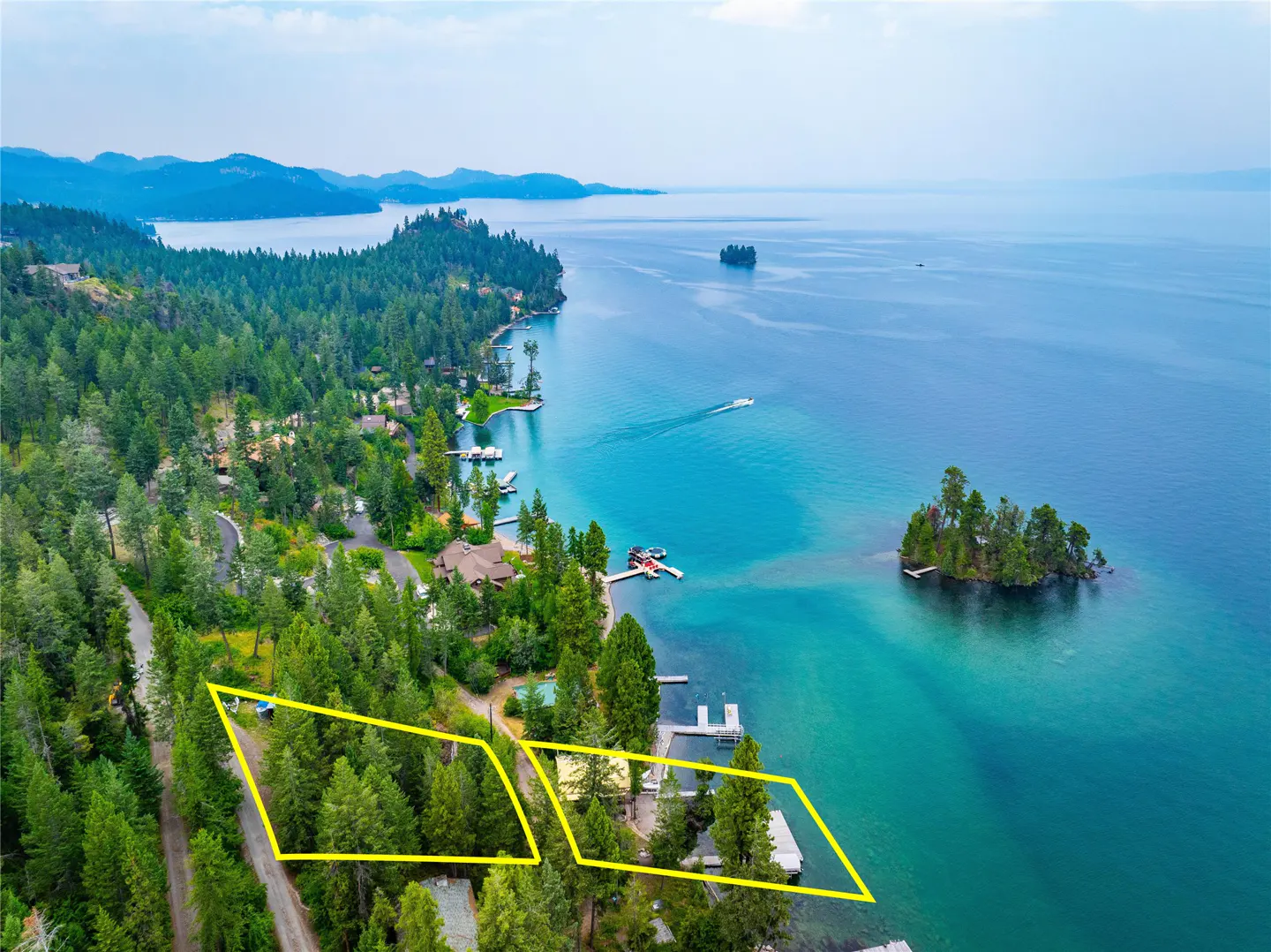 Aerial view of lakefront property with yellow outlines, showcasing land for sale. Blue water, green trees, and distant mountains.
