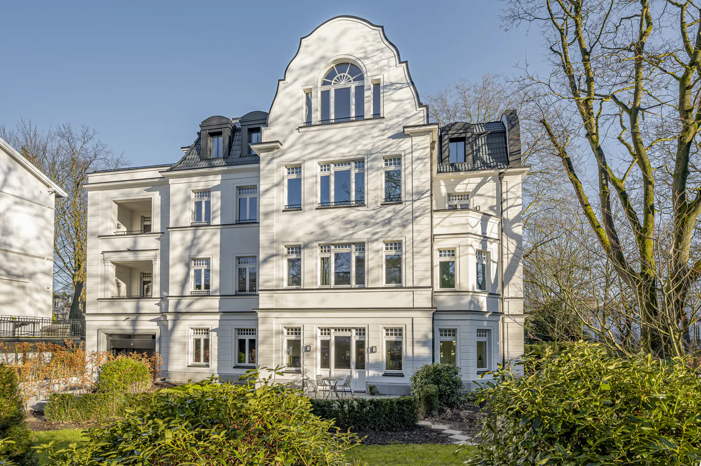 Three-story white building with many windows and a black roof, surrounded by green bushes and trees.