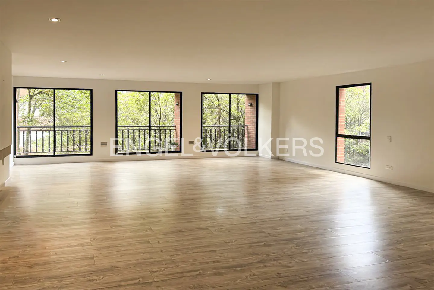 Empty room with light wood floors, white walls, and black-framed windows showing trees and balconies.