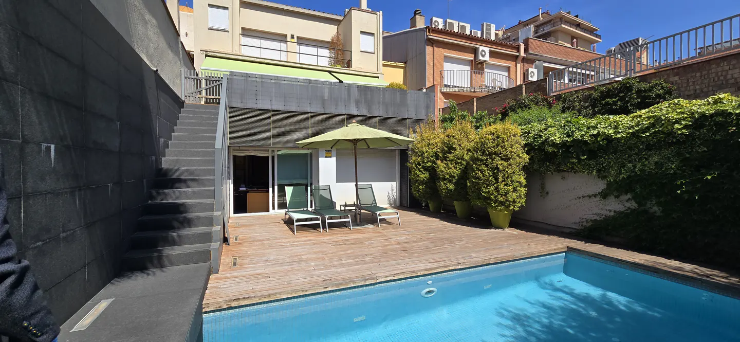 A backyard with a pool, wooden deck, two chairs, and a green umbrella. Stairs lead to the upper level.