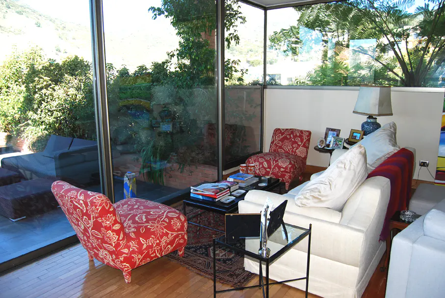 Living room with floor-to-ceiling windows, red floral chairs, white sofas, and a view of lush greenery.