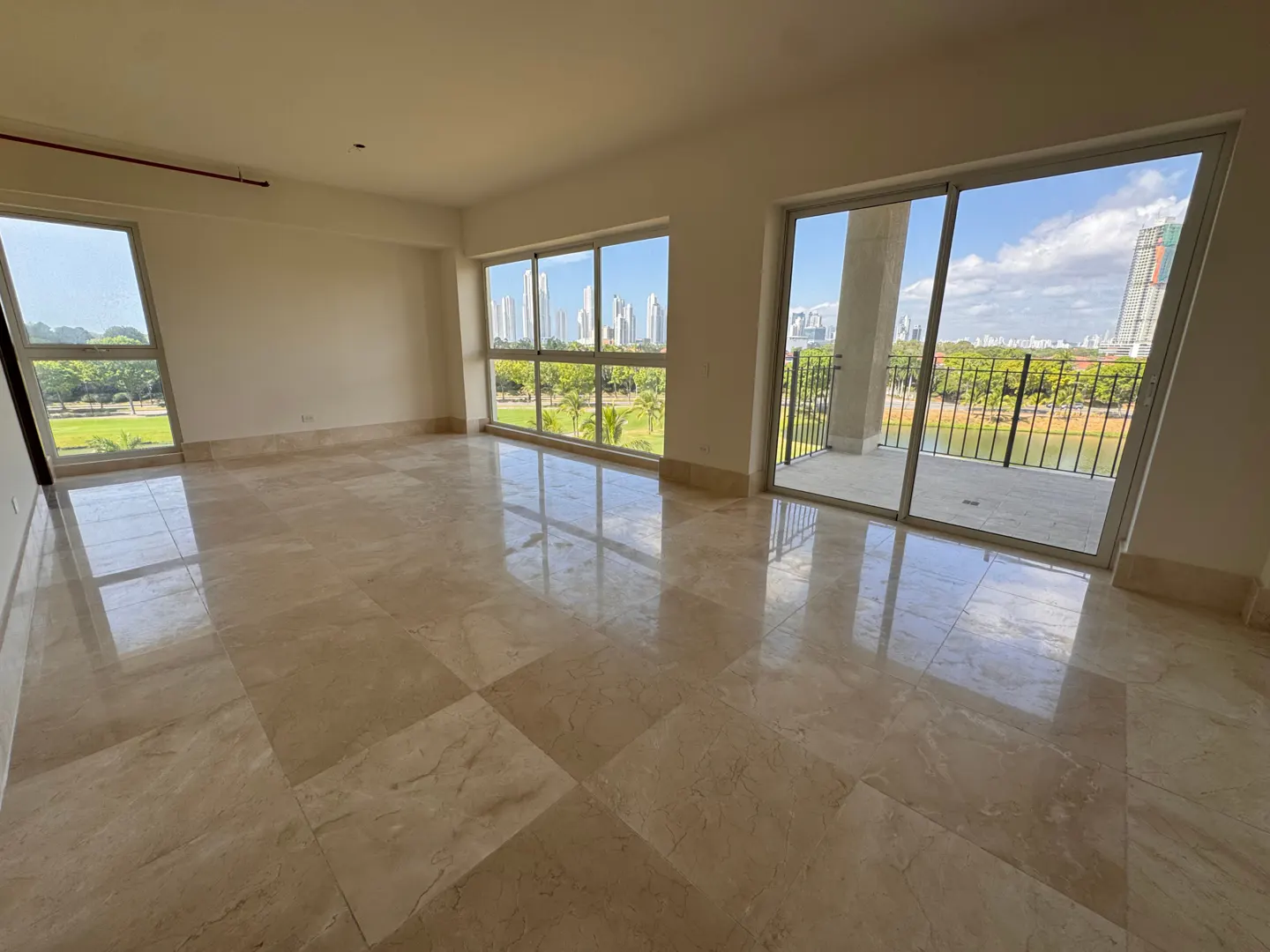 Bright, empty room with beige marble floors and large windows. Balcony with black railing overlooks green space and city skyline.