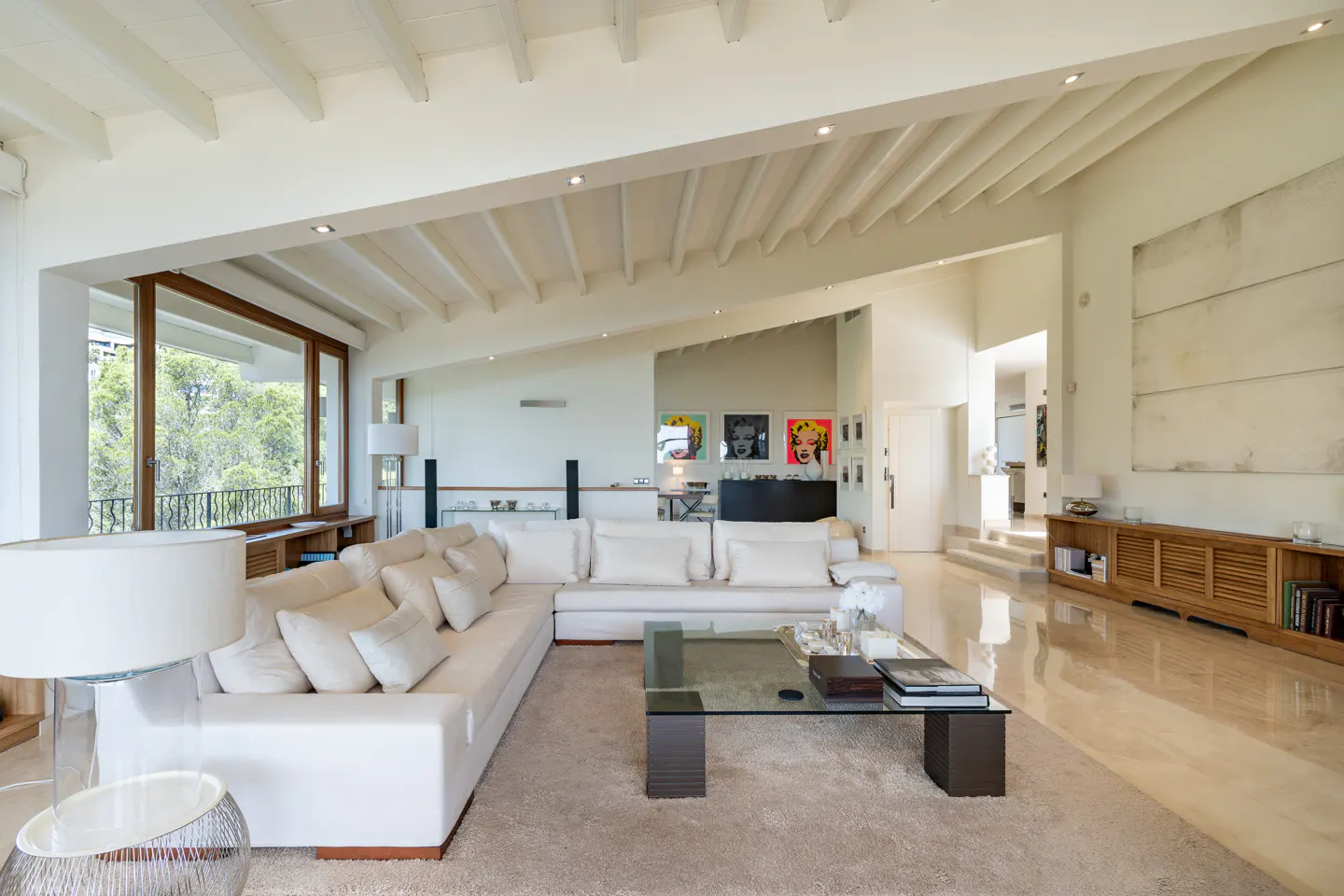 Bright, airy living room with white walls, exposed beam ceiling, and large windows. A white sectional sofa faces a glass coffee table on a beige rug.