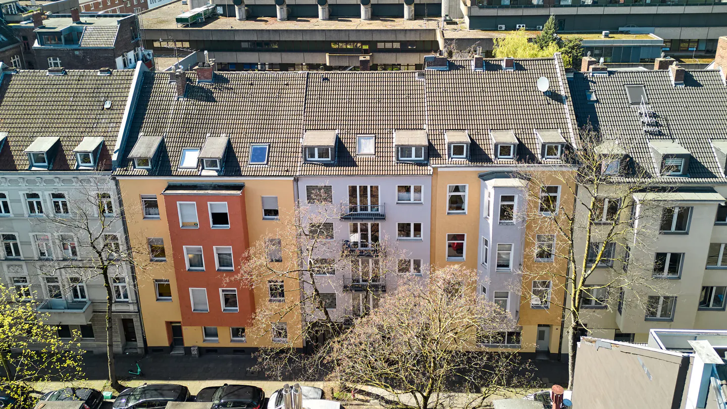Aerial view of a row of colorful urban apartment buildings with tiled roofs and bare trees in front on a sunny day.