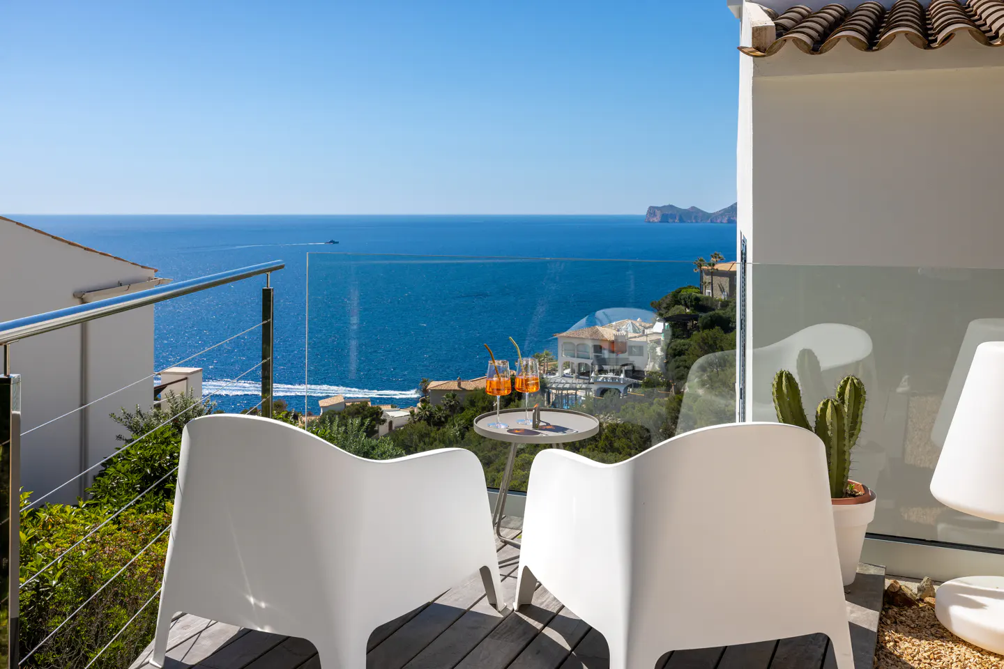 Balcony view with two white chairs, a table with drinks, and a cactus overlooking the blue ocean.