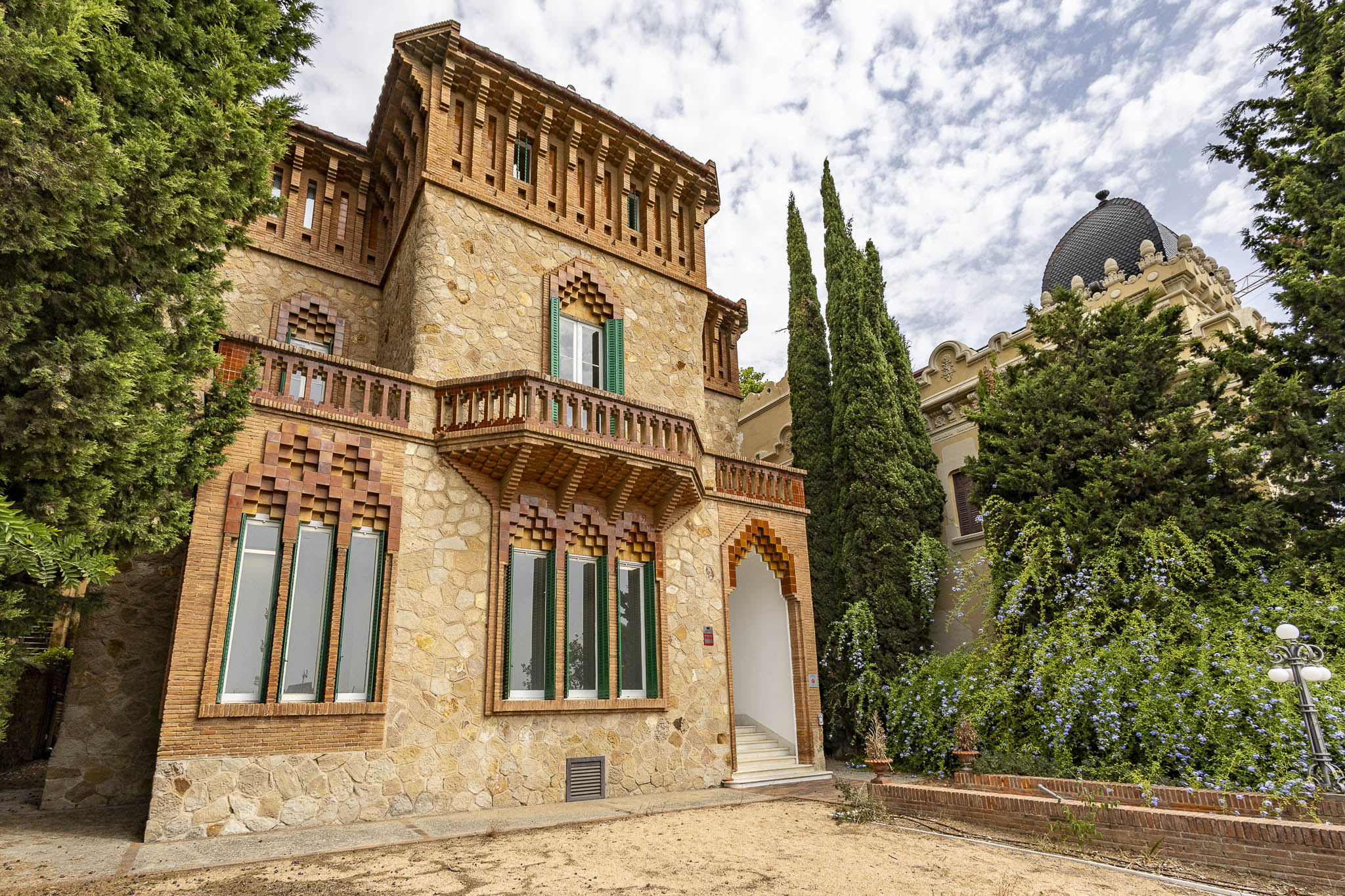 A historic stone mansion with brick detailing, green shutters, a balcony, and tall cypress trees under a cloudy sky.