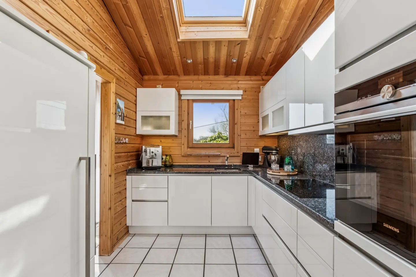 A modern kitchen with wood walls and white cabinets, black granite countertops, and a white tiled floor. A window and skylight provide natural light.