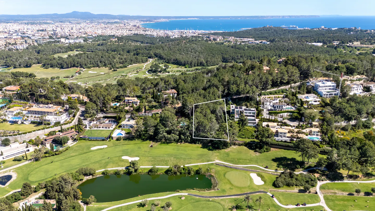Aerial view of a wooded lot outlined in white, nestled in a green golf course near luxury homes, with a city and ocean in the background.