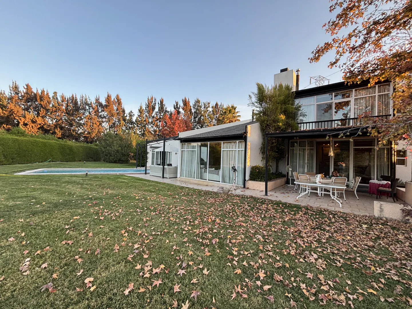 Two-story white house with a patio, table, and chairs. A pool is visible in the background, and leaves are scattered on the lawn.