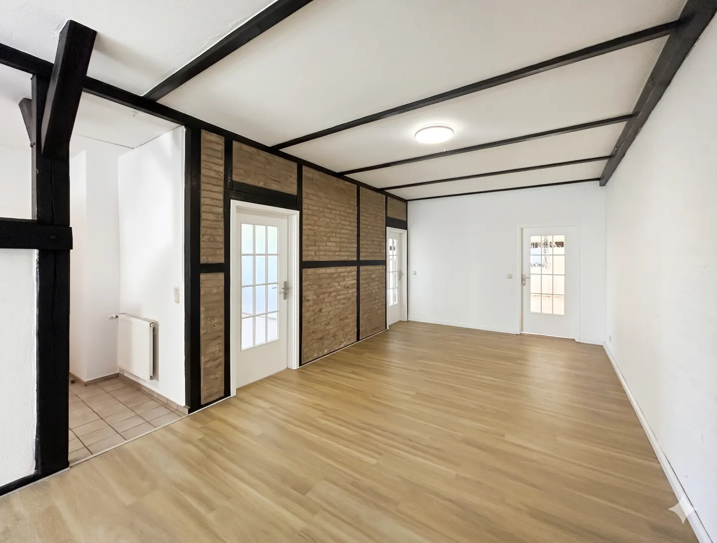 Bright, empty room with wood floors, white walls, and dark wood beams on the ceiling. Two white doors with glass panes are visible.