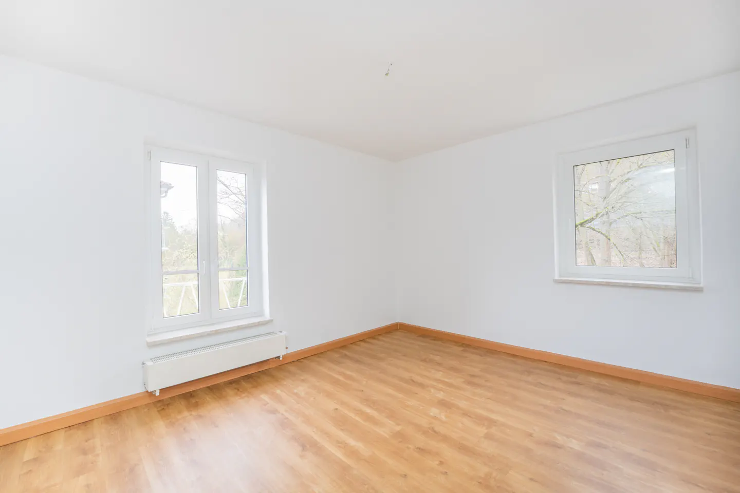 Bright, empty room with light wood floors, white walls, and two white-framed windows showing trees outside.