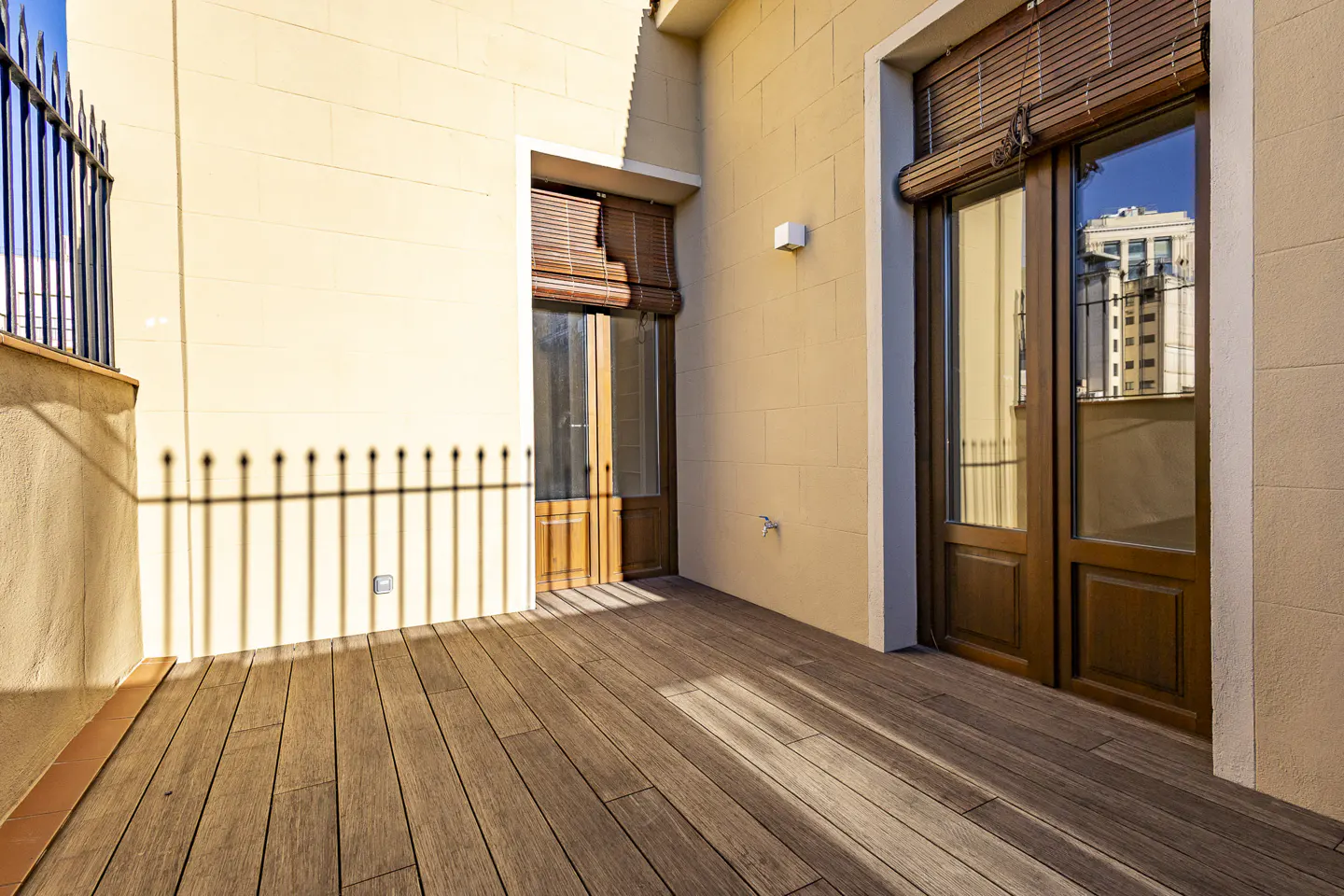 Outdoor patio with wooden floor, beige walls, and two sets of wooden doors with bamboo blinds. Shadows of a black iron fence are cast on the wall.