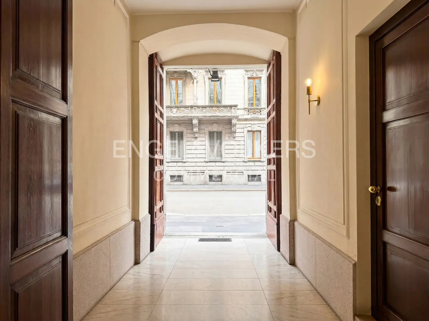 Hallway with marble floors and cream walls leads to open doors revealing a city street with a building facade.