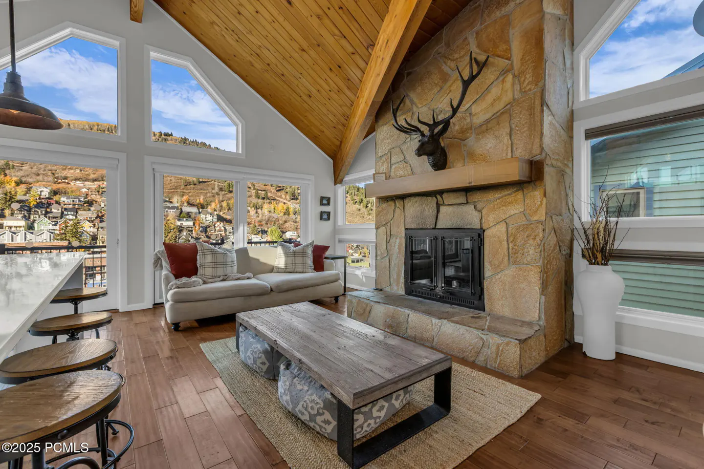 Living room with wood floors, stone fireplace with deer head, and large windows overlooking a hillside town.