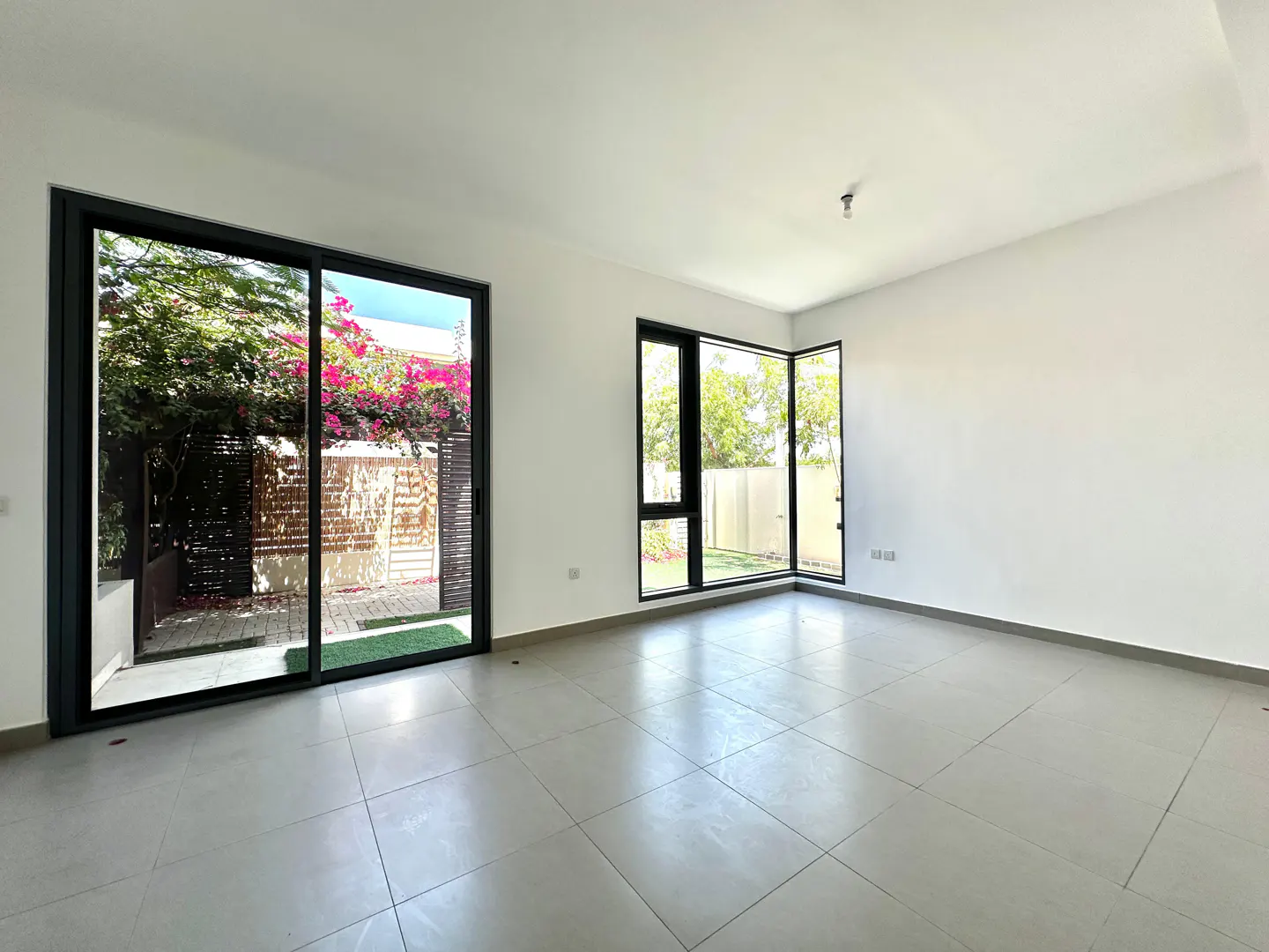 Bright, empty room with white walls, gray tile floor, and black-framed windows. Outside, greenery and pink flowers are visible.