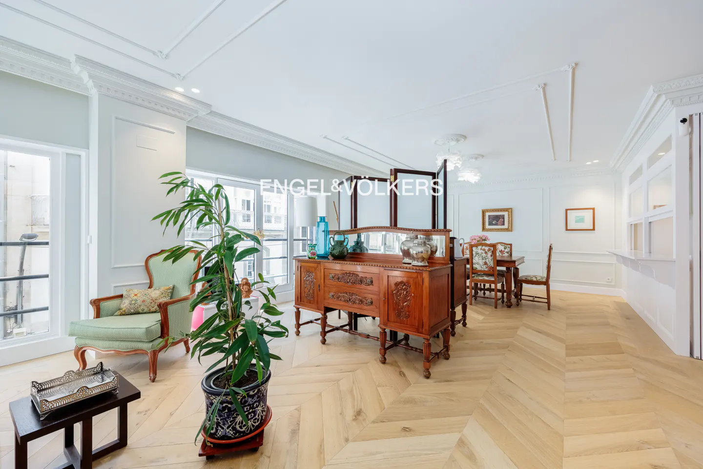Bright living room with herringbone wood floors, antique wood buffet, green chair, and potted plant. White walls and trim.
