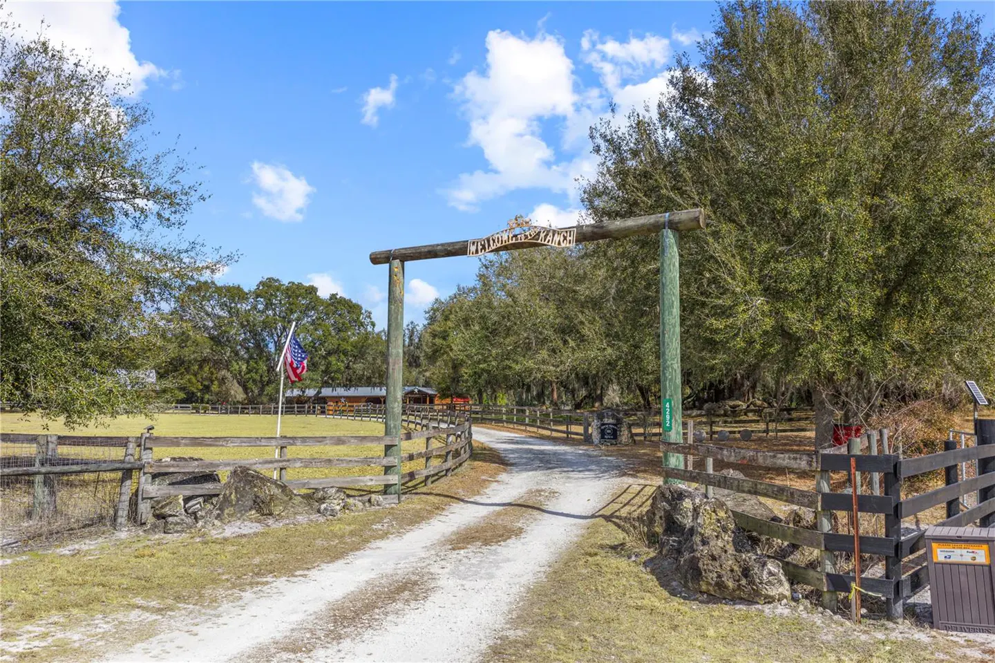 Ranch entrance with wooden archway reading "Welcome to the Ranch," dirt road, wooden fence, and American flag on a sunny day.