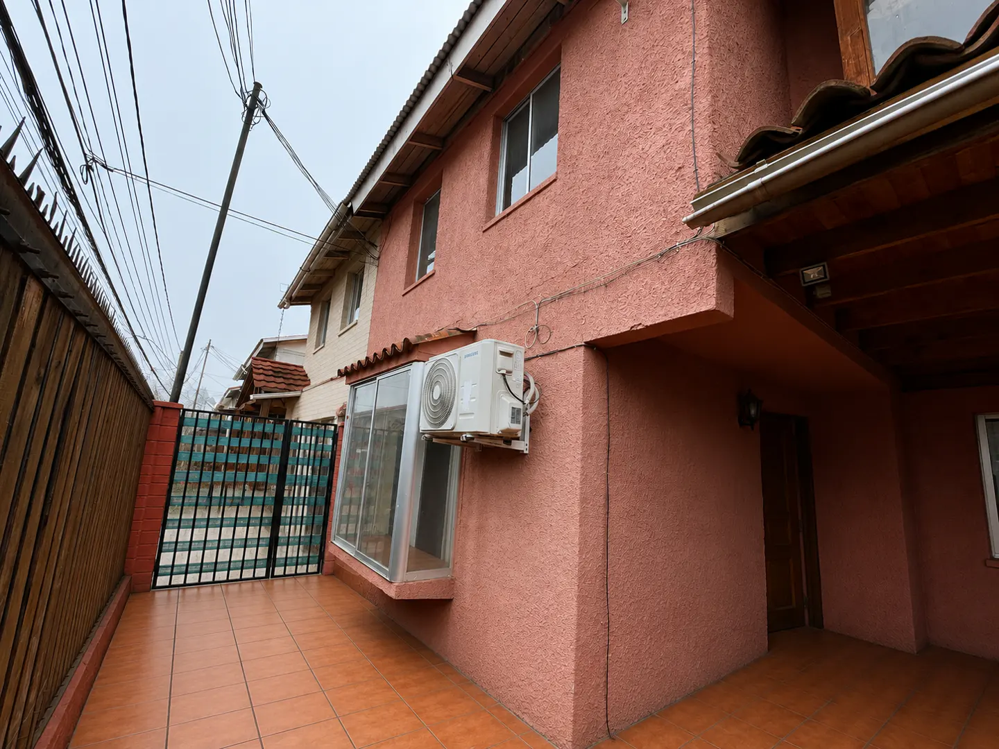 Exterior view of a two-story pink house with a tiled patio, a window, and an air conditioning unit. A wooden fence and a metal gate are visible.