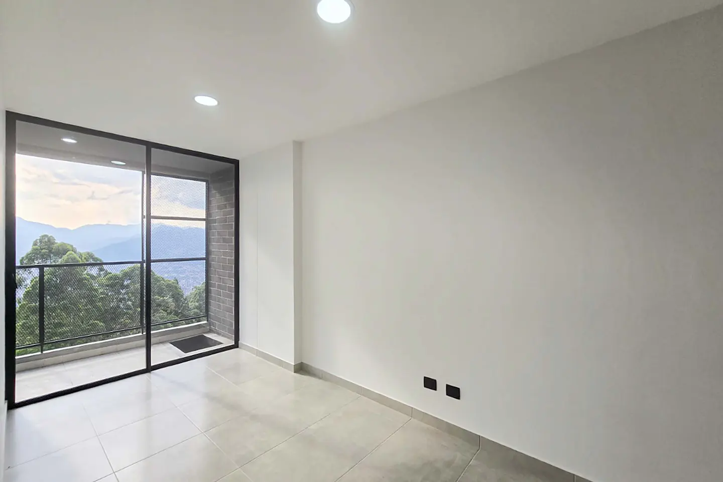 Bright, empty room with gray tile floor and white walls. Black-framed sliding glass doors open to a balcony with mountain views.