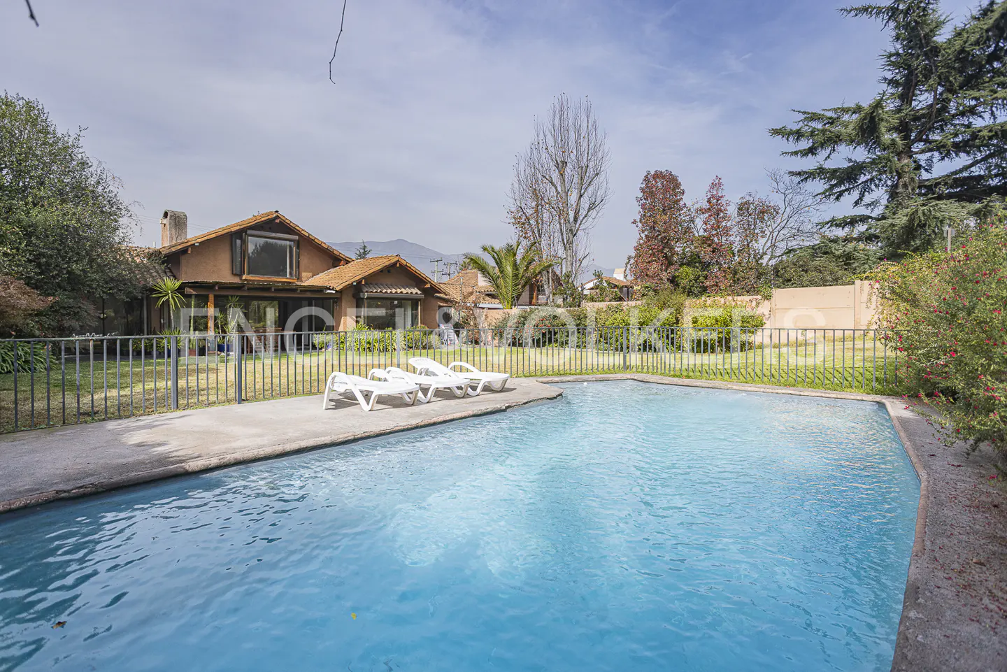 A backyard with a blue swimming pool, white lounge chairs, a brown house, and green trees.