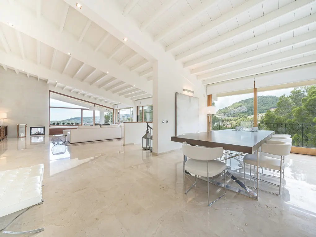Bright, open-plan living space with white walls, wooden ceiling beams, and marble floors. A dining table with white chairs sits near a large window with a green view.