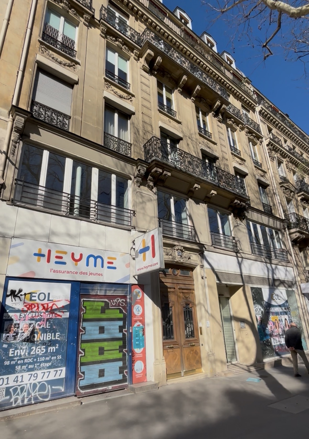 A Parisian building with ornate balconies and windows. Street-level shops have colorful signage and graffiti. A man walks on the sidewalk.