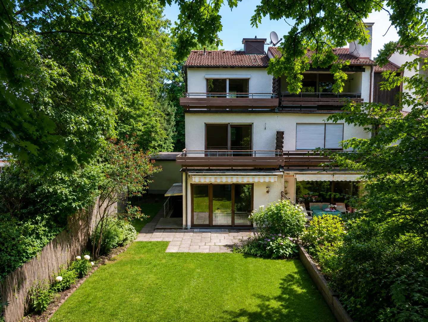 Rear view of a white three-story house with balconies, a stone patio, and a lush green backyard surrounded by trees.