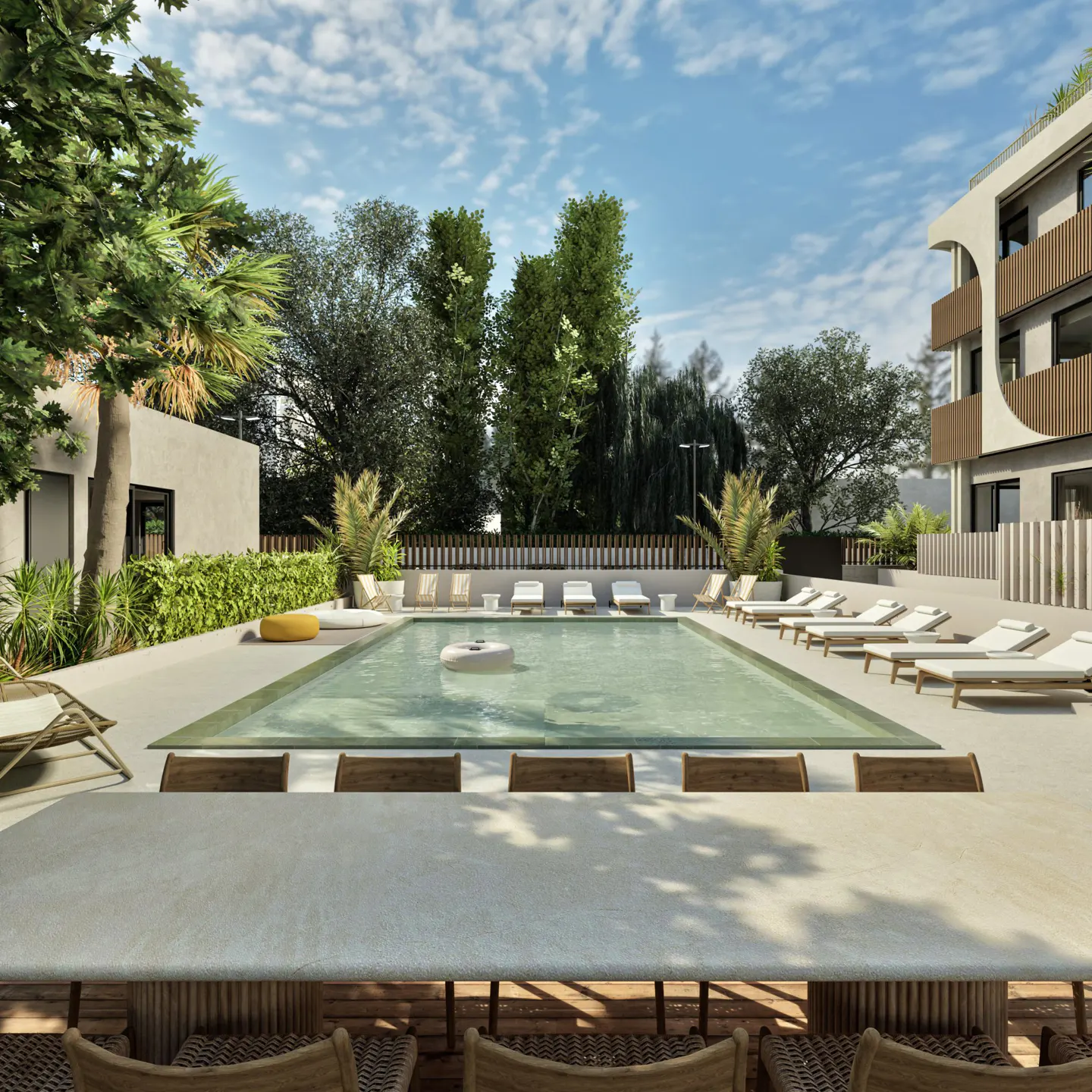 A modern swimming pool area with lounge chairs, a long dining table, and lush trees under a bright blue sky.
