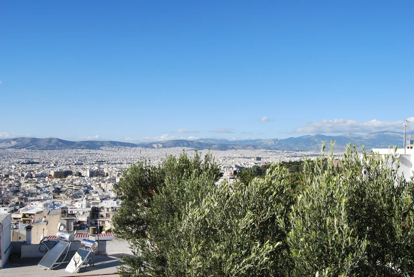 Cityscape view of Athens, Greece, under a blue sky. Green trees in the foreground, mountains in the distance. Solar panels on a rooftop.