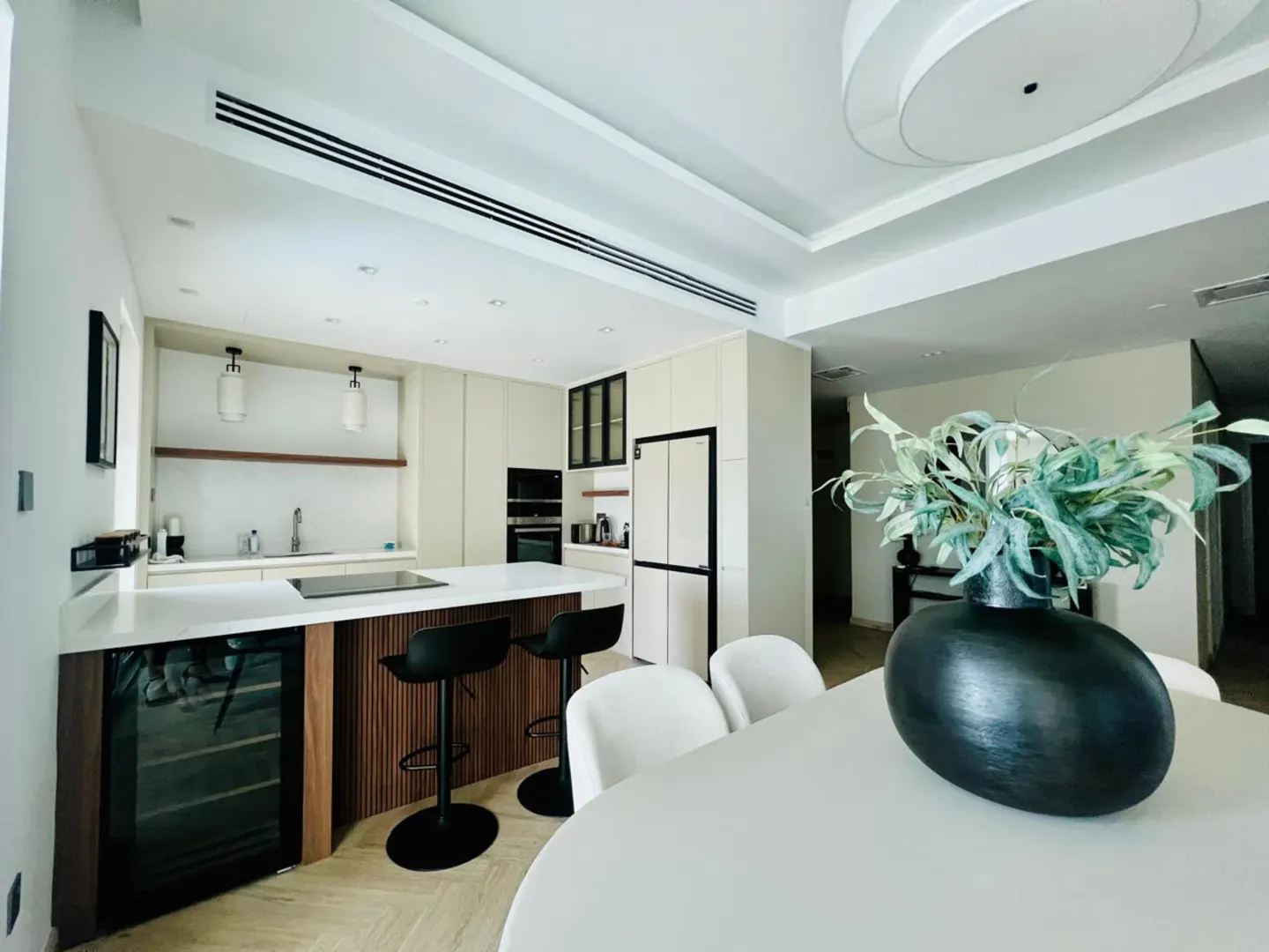 A modern, bright kitchen with white cabinets, a wood-paneled island with black stools, and a dining table with a black vase.